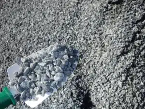 Shovel holding grey crushed stone, with a large pile of aggregate.